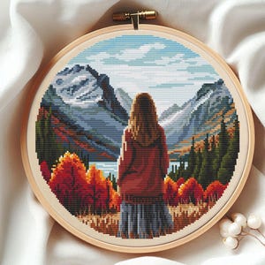 May include: A circular cross-stitch embroidery piece in a wooden hoop featuring a person with long hair wearing a red sweater and a gray skirt, standing before a landscape of autumn trees, a blue lake, and snow-capped mountains under a cloudy sky.