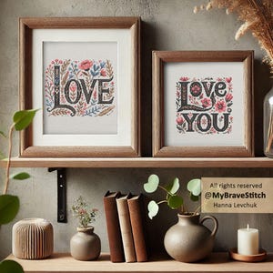 May include: Two framed cross-stitch artworks with the words "LOVE" and "LOVE YOU" in a floral design. The frames are wooden, and the artwork features red, pink, and blue floral accents. The pieces are displayed on a wooden shelf.