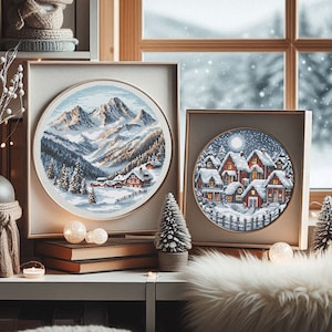 Puede incluir: Dos diseños de bordado de punto de cruz enmarcados. El primer diseño presenta un paisaje montañoso nevado con un pequeño pueblo en la base. El segundo diseño presenta un pueblo nevado con casas y una luna en el cielo.
