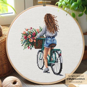 Könnte beinhalten: Kreuzstich-Stickerei in einem Holzreifen zeigt eine Person auf einem teal-farbenen Fahrrad mit einem Blumenkorb. Die Person trägt ein graues Oberteil und Jeansshorts. Die Blumen sind rosa, rot und pfirsichfarben. Der Hintergrund ist weiß.