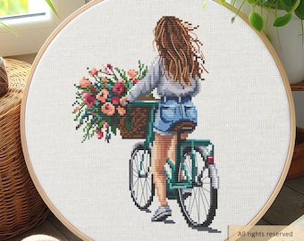 May include: A cross-stitch embroidery in a round wooden hoop depicting a person with long brown hair riding a green bicycle. The bicycle features a brown basket overflowing with pink, orange, and green flowers. The person wears a gray top and blue shorts.