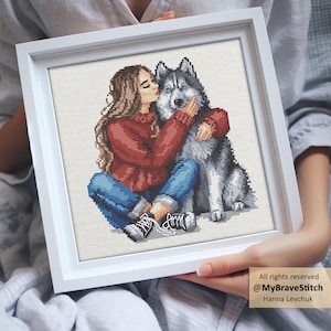 May include: A framed cross-stitch artwork shows a person hugging a husky dog. The person is wearing a red sweater and blue jeans, and the dog has grey and white fur. The artwork is in a white frame and held by a person.