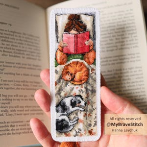 Puede incluir: Un marcapáginas rectangular de tela con un diseño de punto de cruz de una persona de pelo castaño leyendo un libro rojo, un gato naranja enroscado y un perro blanco y negro tumbado sobre una manta texturizada, con bordes cosidos en blanco.