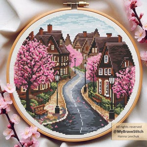 Puede incluir: Un bordado de punto de cruz de una pintoresca escena de pueblo. La obra presenta casas marrones, una carretera sinuosa y vibrantes cerezos en flor rosas. La pieza está en un bastidor de bordado de madera. El texto en la imagen dice: "All rights reserved @MyBraveStitch Hanna Levchuk."