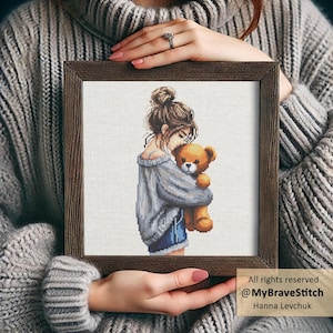 Könnte beinhalten: Kreuzstich-Kunstwerk in einem braunen Holzrahmen. Das Kunstwerk zeigt eine Person, die einen Teddybären umarmt. Die Person trägt einen grauen Pullover und Jeansshorts. Der Teddybär ist braun. Text: All rights reserved @MyBraveStitch Hanna Levchuk.