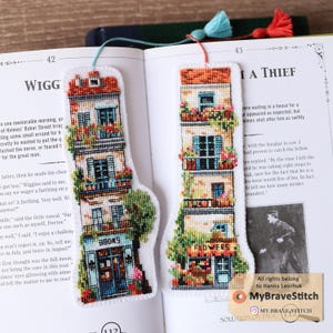 Könnte beinhalten: Zwei gestickte Lesezeichen mit Gebäudedesigns. Eines trägt die Aufschrift "BOOKS" und das andere "FLOWERS". Jedes hat eine Quaste. Die Gebäude haben Balkone mit Blumen und die Dächer sind orange. Die Lesezeichen liegen auf einem Buch.