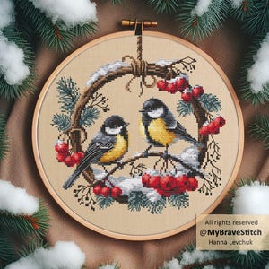 以下が含まれることがあります： 赤い実の枝に止まっている2羽の鳥のクロスステッチ刺繍。雪と松の葉で囲まれています。木製の刺繍枠に入っています。「All rights reserved @MyBraveStitch Hanna Levchuk」のテキストが表示されています。