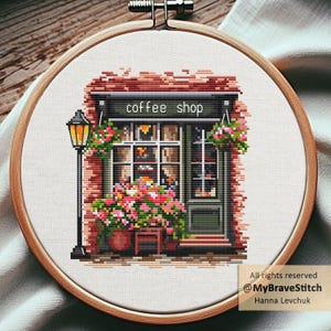 May include: Cross-stitch embroidery of a coffee shop with a brick facade, green door, and window. The shop has a black awning with the words "coffee shop". Pink and red flowers are in pots and hanging from the awning. A black lamppost is on the left.