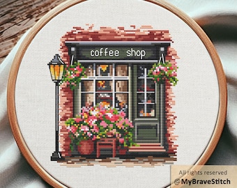 May include: A cross-stitch embroidery in a round wooden hoop depicting a coffee shop storefront with a brick facade, a black lamp post, and colorful flower boxes. The text "coffee shop" is stitched in white above the shop window and door.