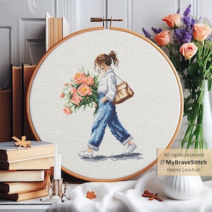 Op de afbeelding: Kruissteekborduurwerk in een houten hoepel toont een persoon die een groot boeket roze bloemen en een bruine tas draagt. De persoon draagt een wit overhemd, een blauwe spijkerbroek en witte sneakers. De tekst "@MyBraveStitch Hanna Levchuk" staat rechtsonder.