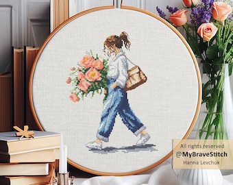 May include: A circular wooden embroidery hoop containing a cross-stitch pattern of a person with brown hair in a ponytail, wearing a white shirt and blue jeans, carrying a large bouquet of pink flowers and a tan shoulder bag on white fabric.