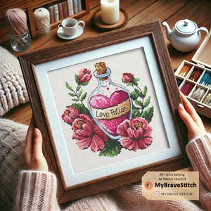 Puede incluir: Un diseño de punto de cruz enmarcado que presenta una botella de vidrio con un tapón de corcho llena de líquido rosa. La botella está etiquetada como "Love Potion" y está rodeada de flores rosas. El diseño está sobre un fondo blanco.