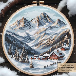 May include: A cross-stitch embroidery of a snowy mountain landscape in a round wooden hoop. The scene features snow-covered mountains, evergreen trees, and a small village with red-roofed houses. The color palette includes shades of blue, white, and brown.