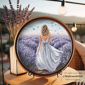 May include: A round, framed cross-stitch artwork depicts a woman in a white dress walking through a lavender field. The artwork is in a brown frame and is displayed on a wooden table. The image includes the text "All rights reserved @MyBraveStitch Hanna Levchuk".