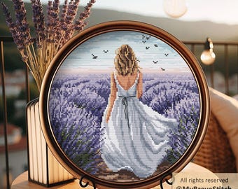 May include: A round, framed cross-stitch artwork depicts a woman in a white dress walking through a lavender field. The artwork is in a brown frame and is displayed on a wooden table. The image includes the text "All rights reserved @MyBraveStitch Hanna Levchuk".