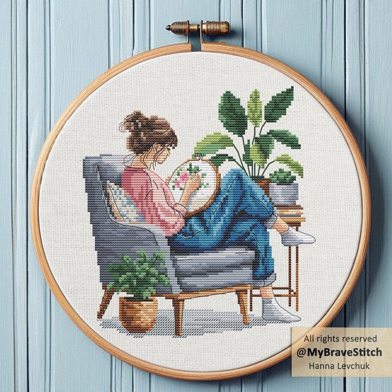 May include: A cross-stitch embroidery hoop featuring a woman seated in a gray armchair, engrossed in her craft. She's wearing a pink shirt and blue jeans. The design includes plants and a floral pattern, with text that reads "All rights reserved @MyBraveStitch Hanna Levchuk."