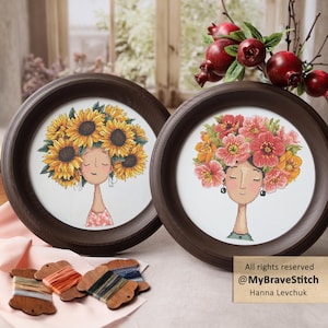 May include: Two circular dark brown wooden frames containing cross-stitch portraits of individuals with closed eyes. One features a person with a large sunflower crown, the other a person with a pink and orange floral crown. Includes thread spools.