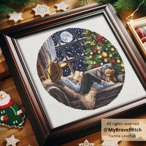 Puede incluir: Arte de punto de cruz enmarcado que representa una escena navide&ntilde;a. Una persona lee junto a una ventana con vistas a una noche nevada y un &aacute;rbol de Navidad decorado. Un osito de peluche est&aacute; en el sof&aacute;. El marco es marr&oacute;n oscuro.