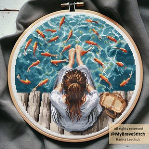 May include: A cross-stitch embroidery in a wooden hoop shows a person relaxing by water with orange fish. The person wears a light blue shirt and has brown hair. The water is teal and the fish are orange. The text reads "All rights reserved @MyBraveStitch Hanna Levchuk".