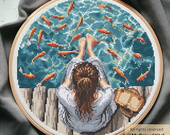 May include: A cross-stitch embroidery in a wooden hoop depicts a person relaxing by a body of water with orange fish. The person is wearing a light blue shirt and has brown hair. The water is teal and the fish are orange.