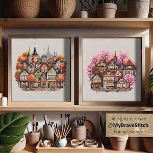 Op de afbeelding: Twee ingelijste kruissteekkunstwerken. Eén toont een kleurrijk dorpsgezicht met herfstbladeren. De andere toont een vergelijkbaar dorp met roze kersenbloesems. Beide bevinden zich in licht houten frames, tentoongesteld op een houten plank met kunstbenodigdheden.