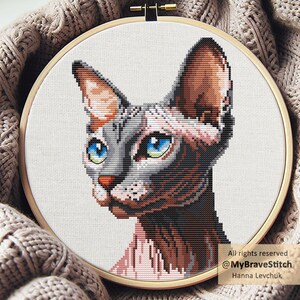Puede incluir: Bordado de punto de cruz de la cara de un gato Sphynx, con tonos grises, marrones y rosas, ojos azules brillantes y una nariz roja. La obra de arte está enmarcada en un aro de madera, sobre un fondo de tela neutra. Texto: Todos los derechos reservados @MyBraveStitch Hanna Levchuk.