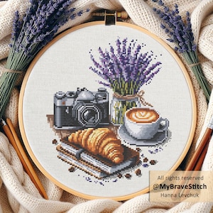 Könnte beinhalten: Kreuzstich-Stickerei in einem Holzrahmen mit einer Kamera, einem Croissant, Kaffee und Lavendel. Das Design umfasst eine schwarz-silberne Kamera, ein goldenes Croissant und eine Tasse Kaffee. Der Text "All rights reserved @MyBraveStitch Hanna Levchuk" ist sichtbar.