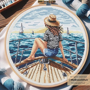 May include: Cross-stitch embroidery in a wooden hoop shows a person on a boat, wearing a straw hat and striped shirt, with a blue ocean and sailboats. The artwork is framed in a wooden hoop.