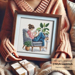 May include: A framed cross-stitch artwork depicts a woman cross-stitching while seated in a gray armchair. The artwork includes potted plants and a small side table. The frame is brown with a white mat. Text in the image reads: All rights reserved @MyBraveStitch Hanna Levchuk.