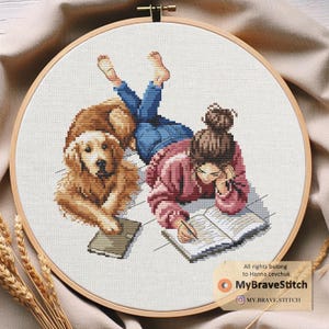 Könnte beinhalten: Kreuzstich-Stickerei in einem Holzreifen. Das Design zeigt eine Person, die auf dem Boden liegt und in ein Buch schreibt, mit einem Golden Retriever-Hund. Die Person trägt einen rosa Pullover und blaue Jeans. Der Hund ist braun. Der Text "MyBraveStitch" ist sichtbar.