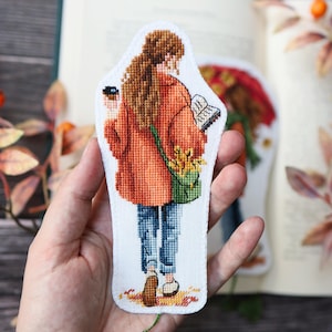 May include: A cross-stitch bookmark featuring a person with long brown hair seen from behind, wearing an orange jacket and blue jeans. The figure carries a green bag with yellow flowers, holds a book, and walks over scattered autumn leaves.