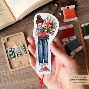 May include: A white fabric bookmark featuring a cross-stitched illustration of a person with a brown bun hairstyle wearing blue denim overalls and red trainers, reading a red book while holding a large bouquet of colourful flowers.