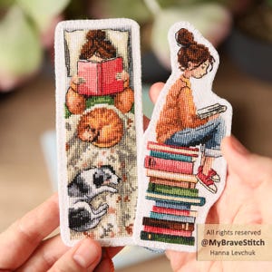 Puede incluir: Dos marcapáginas bordados. Uno presenta a una persona leyendo un libro rojo con un gato y un perro durmiendo. El otro muestra a una persona sentada en una pila de libros, leyendo. Los marcapáginas son blancos con diseños coloridos de punto de cruz.