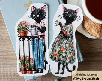 May include: Two embroidered bookmarks featuring black cats. One cat wears a pink shirt and blue pants while watering potted flowers on a balcony. The other cat wears round glasses and a floral dress while holding a book and a small teacup.