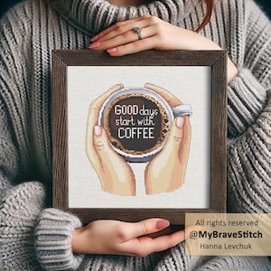 May include: A square cross-stitch embroidery in a dark brown wooden frame featuring a graphic of two hands holding a white mug filled with dark coffee. The text "GOOD days start with COFFEE" is centred within the mug against the dark liquid.
