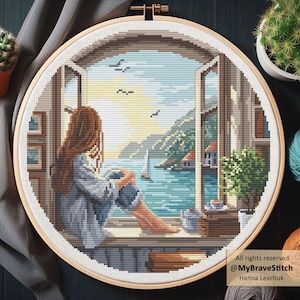 Puede incluir: Un bordado de punto de cruz en un aro de madera representa a una mujer mirando por una ventana una escena costera. La escena incluye un velero, montañas y una taza de café. El bordado está en tonos azules, marrones y verdes.