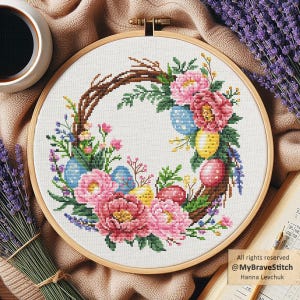 Puede incluir: Un bastidor de bordado de punto de cruz con un diseño de corona de Pascua. La corona está compuesta por peonías rosas, huevos de Pascua azules, amarillos y rojos, y follaje verde, todo sobre un fondo blanco. El bastidor de madera es visible.