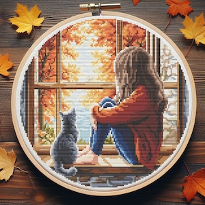 Peut inclure: Broderie au point de croix dans un cercle en bois représentant une personne et un chat regardant par une fenêtre une scène d'automne. La personne porte une chemise rouge et un jean bleu. Le chat est gris. La fenêtre révèle un feuillage d'automne orange et jaune.