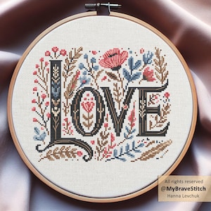 May include: A cross-stitch embroidery hoop featuring the word "LOVE" in black, surrounded by floral and botanical elements. The design includes red, blue, and brown accents, set against a white fabric background. The hoop is made of wood.