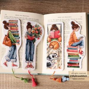 Könnte beinhalten: Vier bestickte Lesezeichen mit Illustrationen von lesenden Personen, mit Bücherstapeln und floralen Elementen. Jedes Lesezeichen hat eine Quaste. Die Lesezeichen sind auf den Seiten eines Buches platziert.