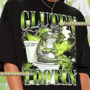 Op de afbeelding: Zwart T-shirt met een groene kikker die op een toilet zit en de tekst "Gluten Got Me Tootin'" in groene en witte letters.