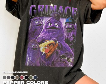 Puede incluir: Una camiseta gris carbón de manga corta con un gran estampado gráfico del personaje morado Grimace en varias poses, incluyendo una con gorra de béisbol y un guante. El texto GRIMACE está impreso en letras moradas grandes arriba.