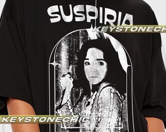 Puede incluir: Camiseta negra con un estampado gráfico blanco de la cara de una mujer con el texto "SUSPIRIA KEYSTONECHIC DO YOU KNOW ANYTHING ABOUT... WITCHES?"