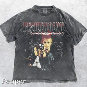 Puede incluir: Camiseta gris oscuro con el texto "RESIDENT EVIL DEAD AIM" en rojo y blanco. El gráfico muestra tres personajes con armas. La camiseta tiene un aspecto vintage y desgastado.