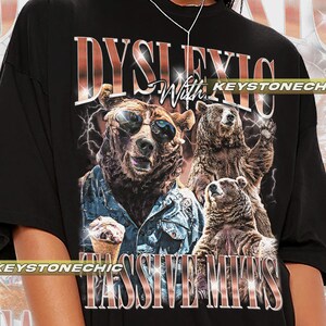 Könnte beinhalten: Schwarzes T-Shirt mit einem grafischen Druck von drei Bären, die Sonnenbrillen tragen und Eis essen. Der Text "Dyslexic With Passive Mits" ist in Pink auf dem Shirt gedruckt.