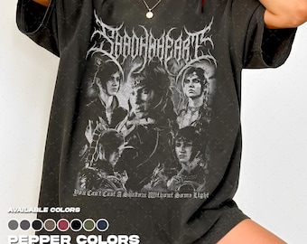 Puede incluir: Una camiseta de manga corta gris carbón con un estampado gráfico de cinco figuras en estilo de fantasía oscura. El texto SHADOWHEART aparece en una fuente estilizada sobre las figuras, con la frase You Can't Cast A Shadow Without Some Light debajo.