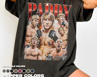 Puede incluir: Una camiseta gris carbón de manga corta con un estampado gráfico de un luchador en varias poses, con el texto PADDY en la parte superior y The Baddy en la parte inferior. El diseño incluye varias imágenes de la misma persona con guantes negros.