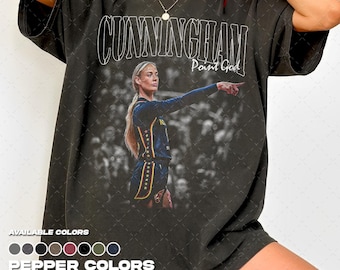 Puede incluir: Una camiseta de manga corta gris carbón con un estampado gráfico de un jugador de baloncesto con uniforme azul oscuro señalando hacia adelante. El texto CUNNINGHAM y Point God está impreso en blanco sobre el jugador. Cuello redondo.