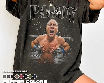 Puede incluir: Una camiseta de manga corta gris carbón con un gran estampado gráfico de un luchador con la boca abierta, usando guantes negros. El texto PADDY PIMBLETT está impreso en una fuente blanca grande sobre la imagen. La camiseta tiene un aspecto desgastado.