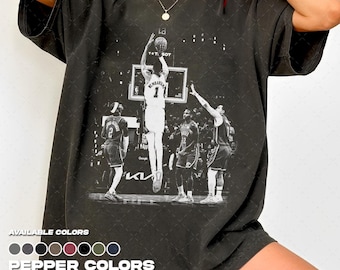 Puede incluir: Camiseta gris oscuro con una imagen en blanco y negro de una acción de baloncesto. La imagen muestra a un jugador lanzando a canasta, con otros jugadores al fondo. El texto "LA" y "T&T SOT" son visibles. La camiseta también muestra "AVAILABLE COLORS" y "PEPPER COLORS".